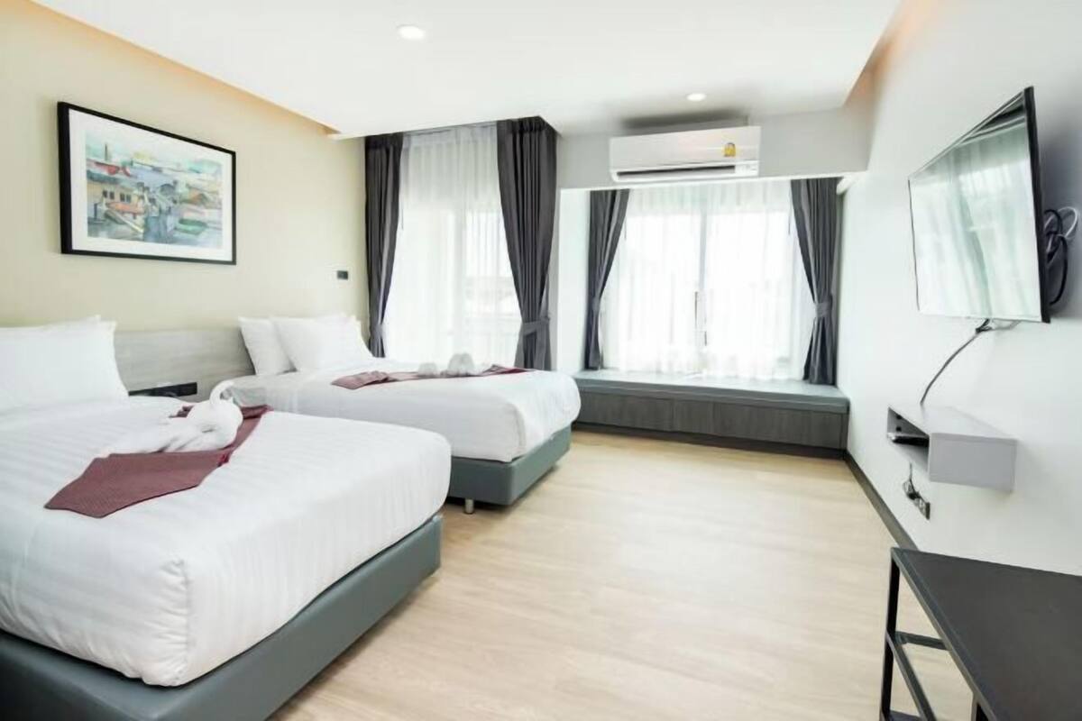 A hotel room with two beds, a large window with curtains, an air conditioning unit, and a flat-screen TV mounted on the wall.