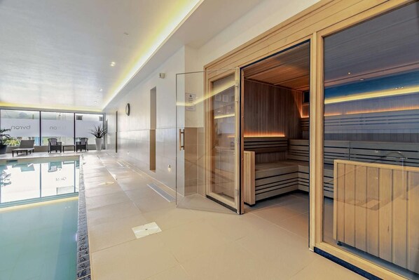 A modern indoor pool area with a glass enclosure, a sauna, and a seating area with a view of the pool.