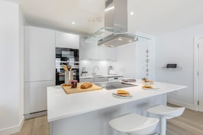 A modern kitchen with white cabinetry, a glass island with stools, a built-in oven, and a stainless steel range hood.
