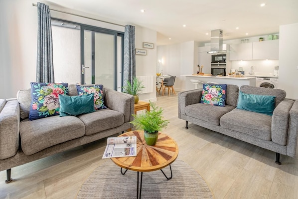 A modern living room with two grey sofas, a coffee table, and a dining area in the background.