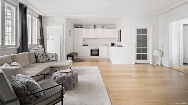 Una sala de estar moderna con un sofá, un reposapiés y una zona de comedor con una cocina al fondo.