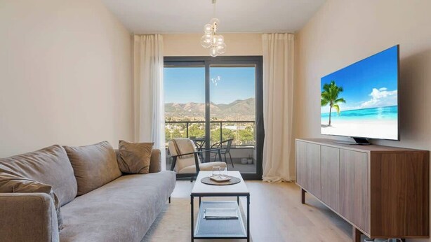 A modern living room with a sofa, coffee table, and TV on a wooden cabinet. A balcony with a view of mountains and a palm tree is visible through the glass doors. - Holiday Home with Pool by Costarentals - Ref 251 (Las Lagunas de Mijas)