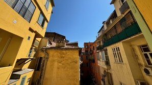 Un stretto vicolo tra edifici con balconi, un cielo azzurro limpido e una finestra con zanzariera ben visibile.