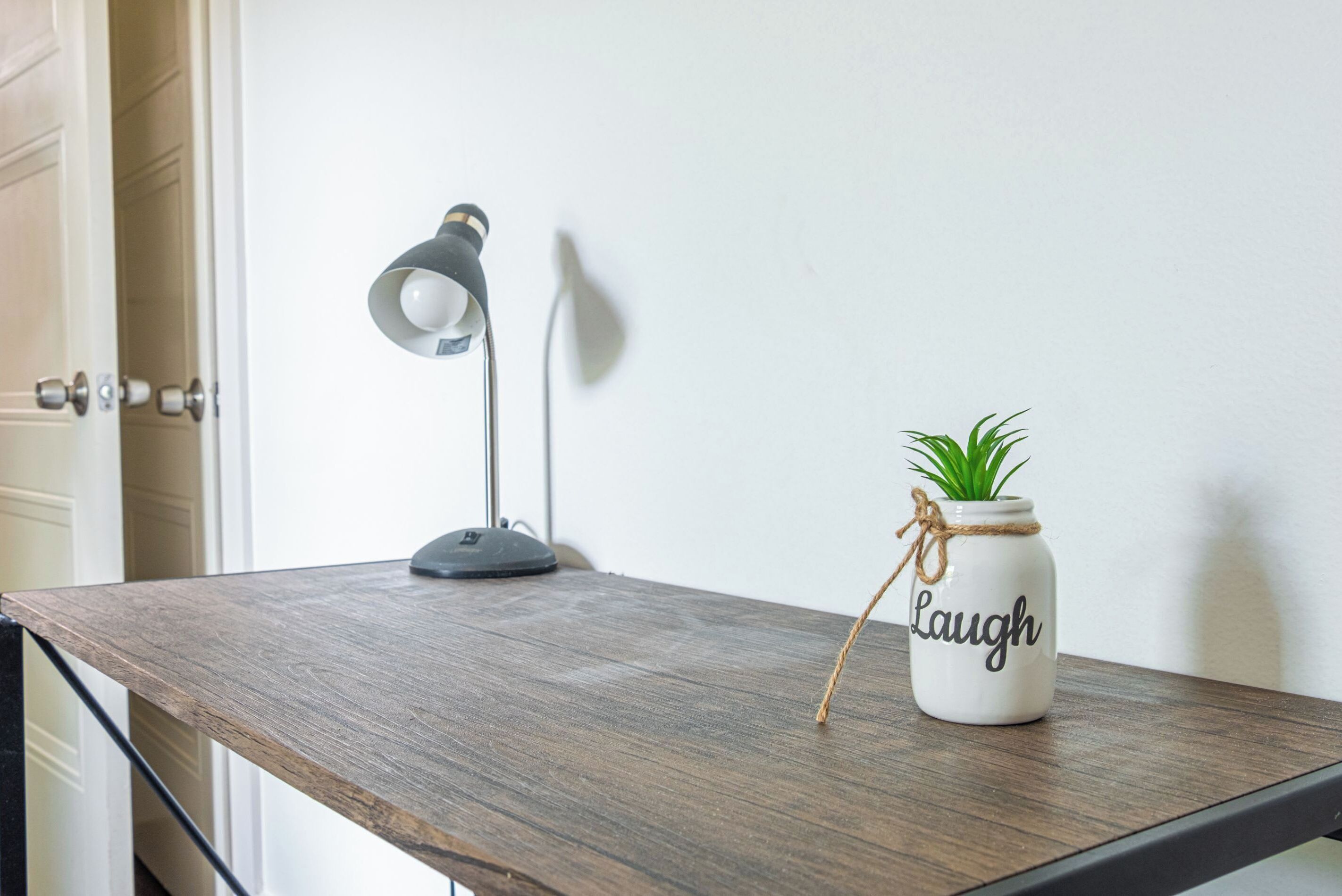 Une table en bois, avec une lampe de bureau et une plante en pot dans un bocal sur lequel est écrit le mot « Rire ».
