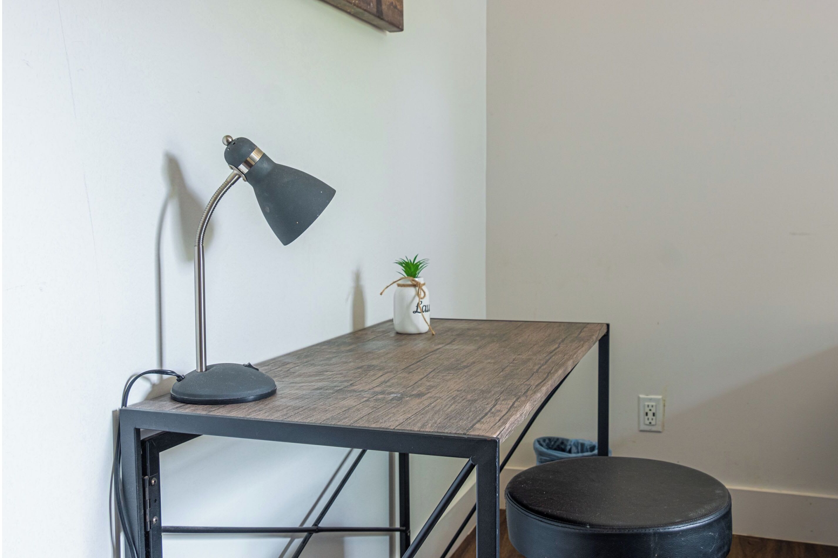 Um pequeno espaço de trabalho com uma escrivaninha, uma luminária, um vaso de plantas e um banquinho.