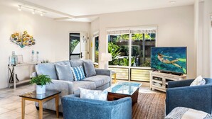 A modern living room with a grey sofa, blue armchairs, a glass coffee table, and a television displaying a swimming scene.