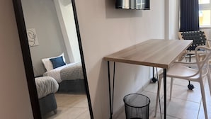 A modern room with a wooden table, white chairs, a bed with a blue pillow, a black trash bin, and a wall-mounted TV. - Phaedrus Living: Hoopoe Residencies 105 (Nicosia)