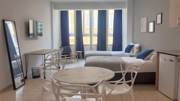 A modern hotel room with a bed, a dining table, chairs, a small kitchen area, and large windows with curtains. - Phaedrus Living: Hoopoe Residencies 105 (Nicosia)