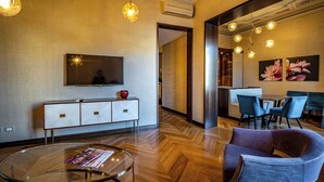 A modern living room with a glass coffee table, a flat-screen TV, a wooden herringbone floor, and a dining area with blue chairs and a wooden table. - YID SMN LUX (Florence)