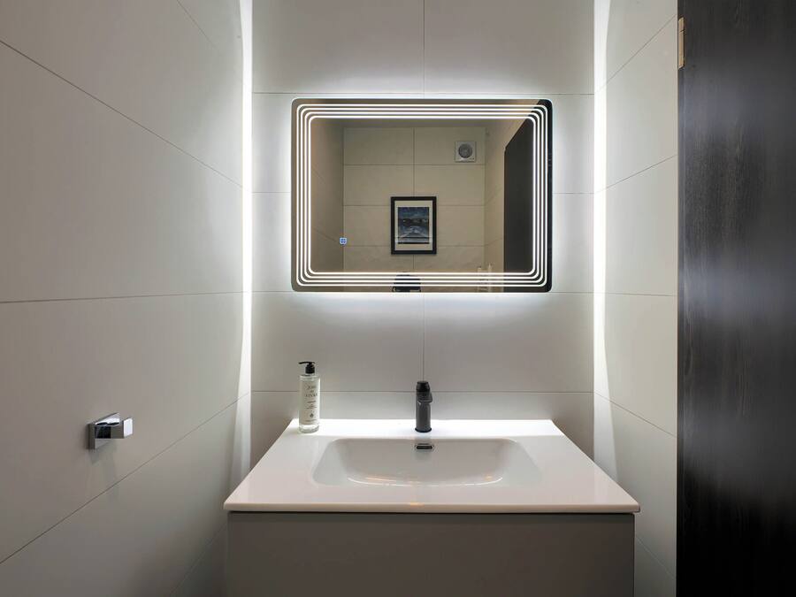 Un baño moderno con un lavamanos blanco, un espejo con marco iluminado y una botella de jabón para manos.