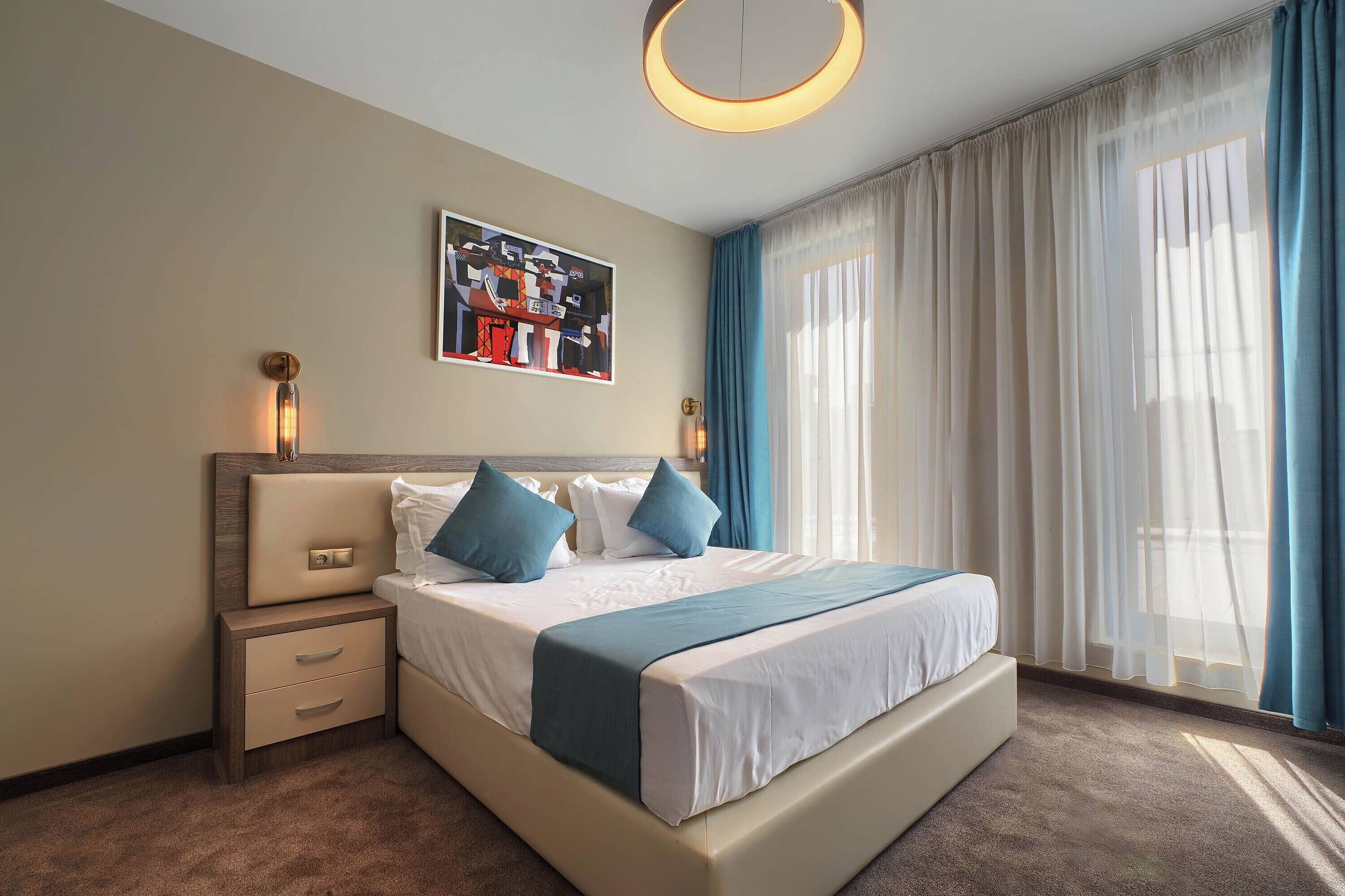 A modern hotel room with a large bed, a bedside table, a nightstand, a wall-mounted light, and a window with sheer curtains.
