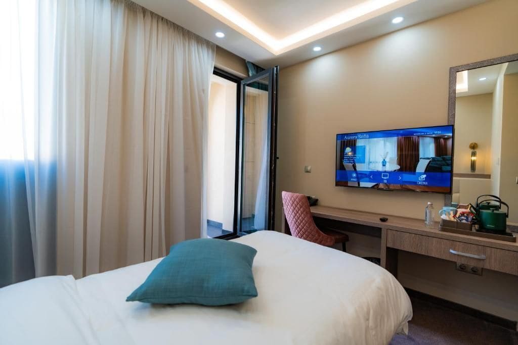 Habitación de hotel con cama, escritorio, televisión y ventana con cortinas.