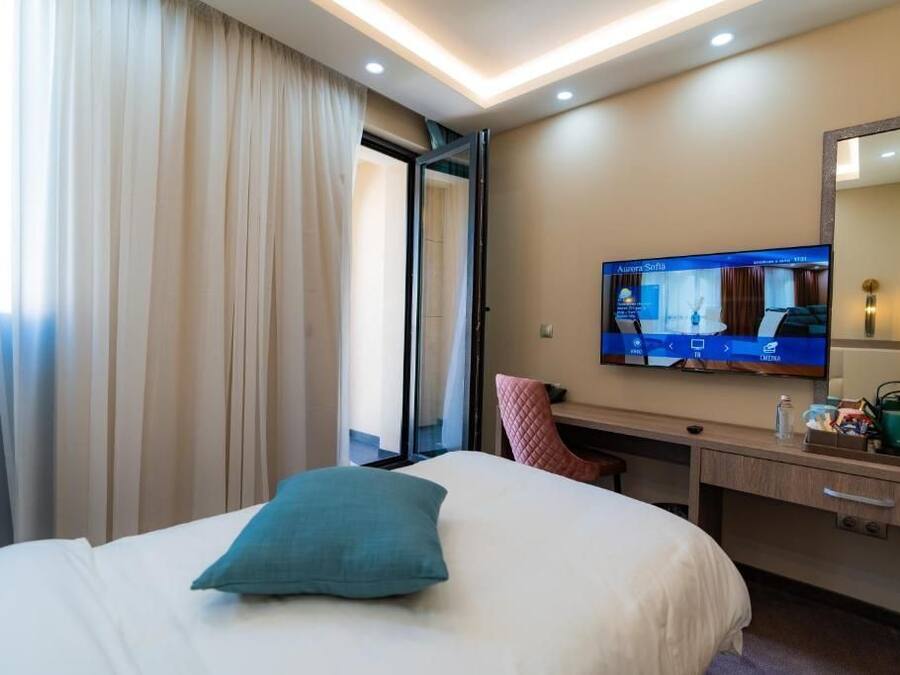 Habitación de hotel con cama, escritorio, televisión y ventana con cortinas.
