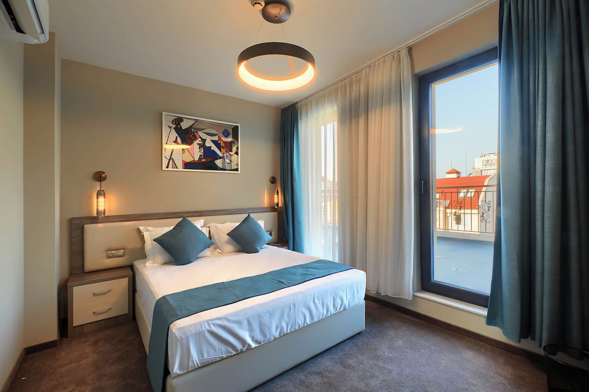 A hotel room with a large bed, a nightstand, a wall-mounted painting, a ceiling light fixture, and a window with curtains.