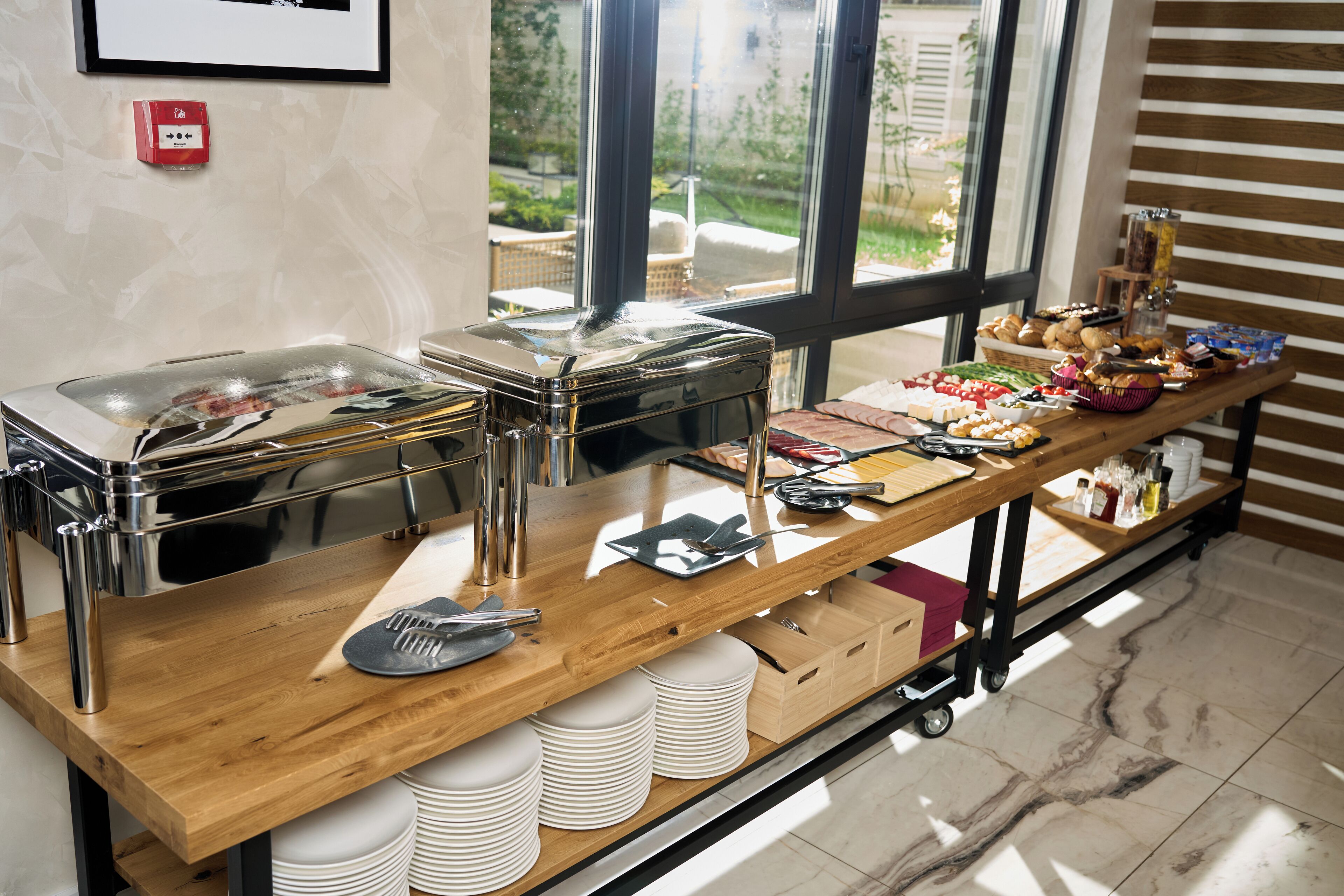A buffet setup with multiple food stations, a wooden table, and a view of the outdoors through large windows.