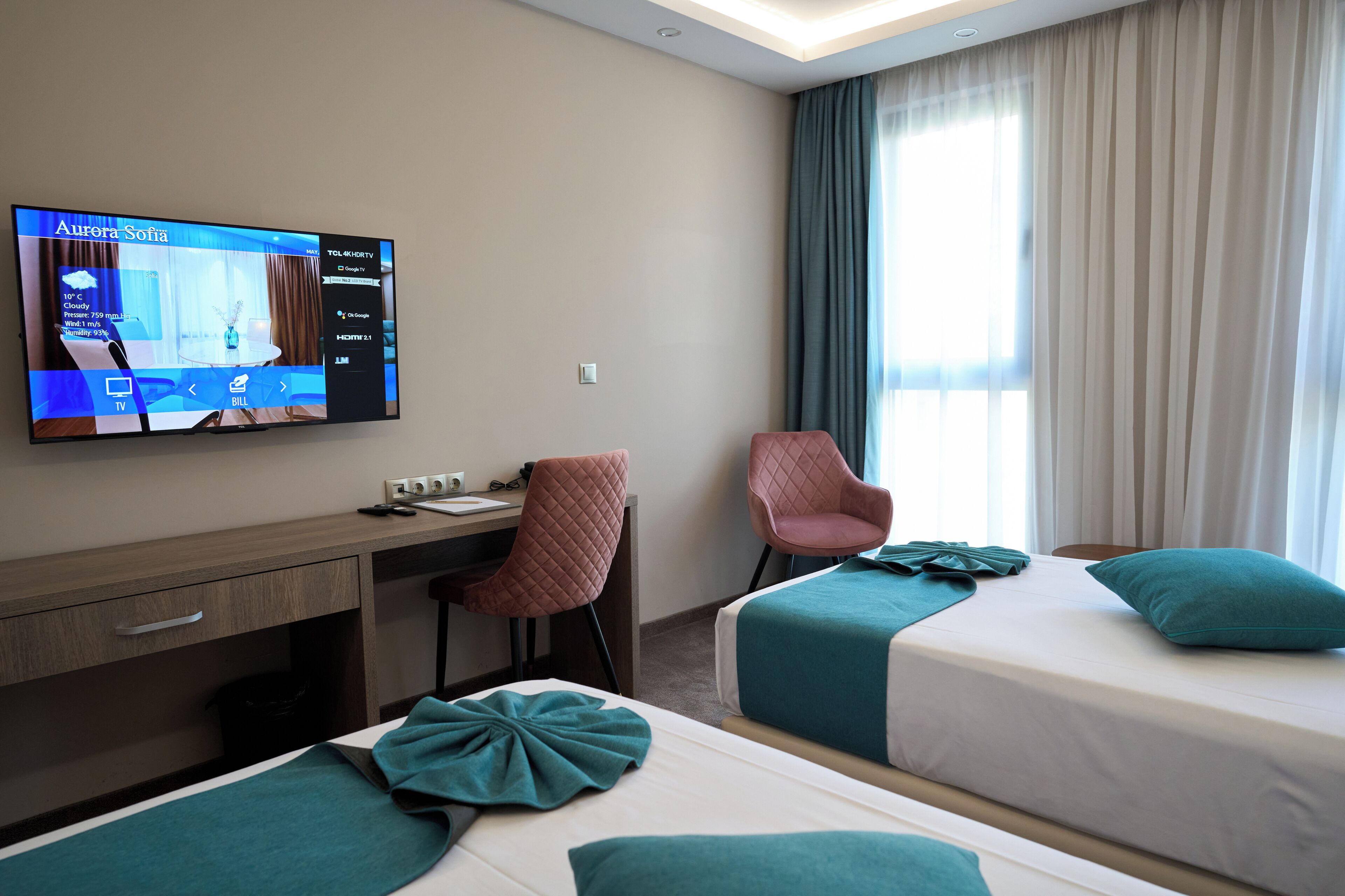 A hotel room with a TV displaying room details, a desk with a chair, two beds with teal bedding, and a large window with curtains.
