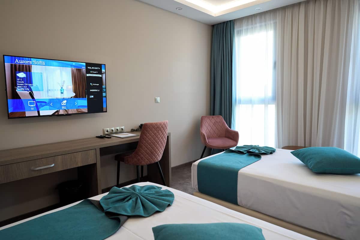 A hotel room with a TV displaying room details, a desk with a chair, two beds with teal bedding, and a large window with curtains.