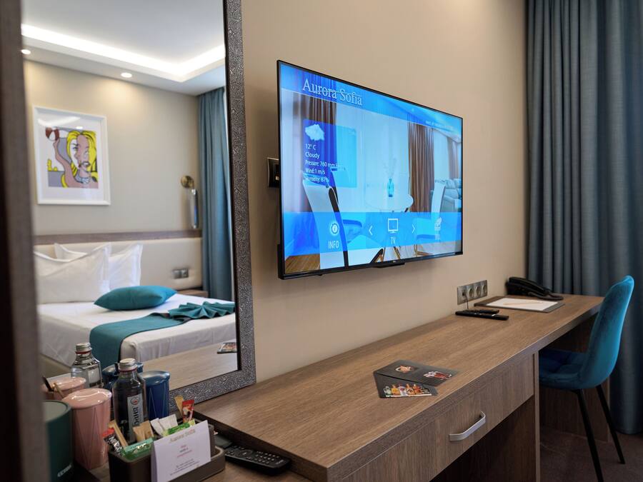 A hotel room with a large flat-screen TV mounted on the wall displaying weather information for 'Aurora Sofia'. There is a wooden desk with a chair, a bed with a blue pillow, and a framed picture on the wall.
