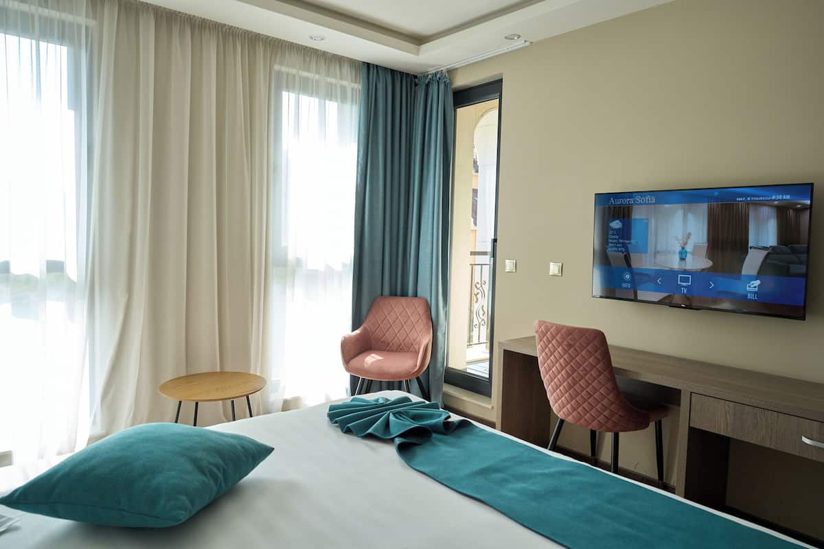 A hotel room with a bed, two chairs, a TV, and a view of the outside.