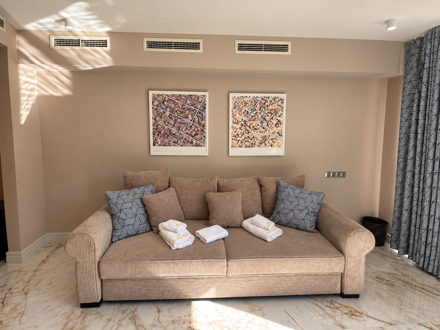 Un sofá tapizado en tono beige con almohadones estampados, dos cuadros abstractos enmarcados en la pared y un suelo de mármol.