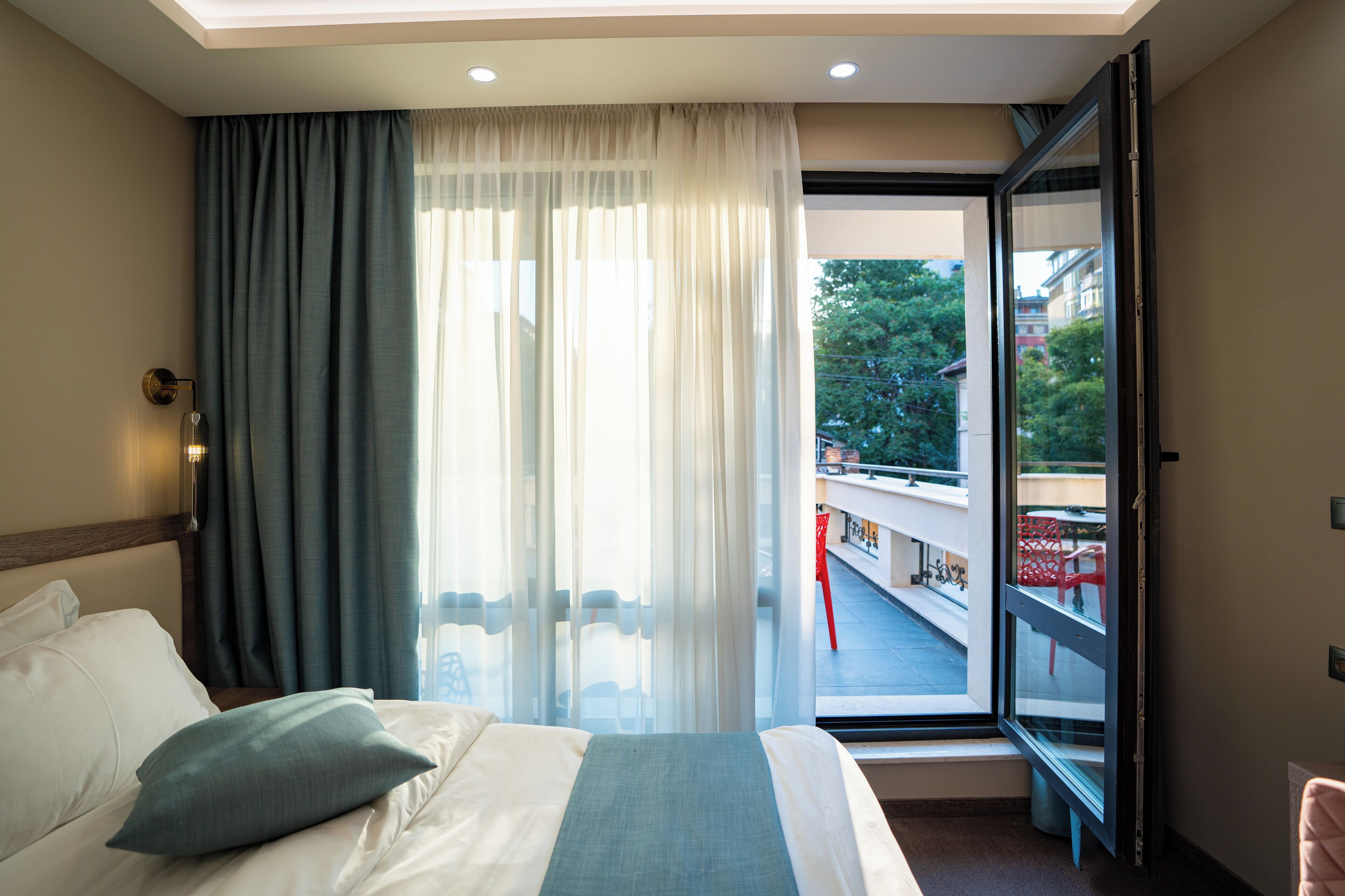 Una camera d'albergo con un letto, una finestra con tende, un balcone con sedie rosse e vista su edifici e alberi.