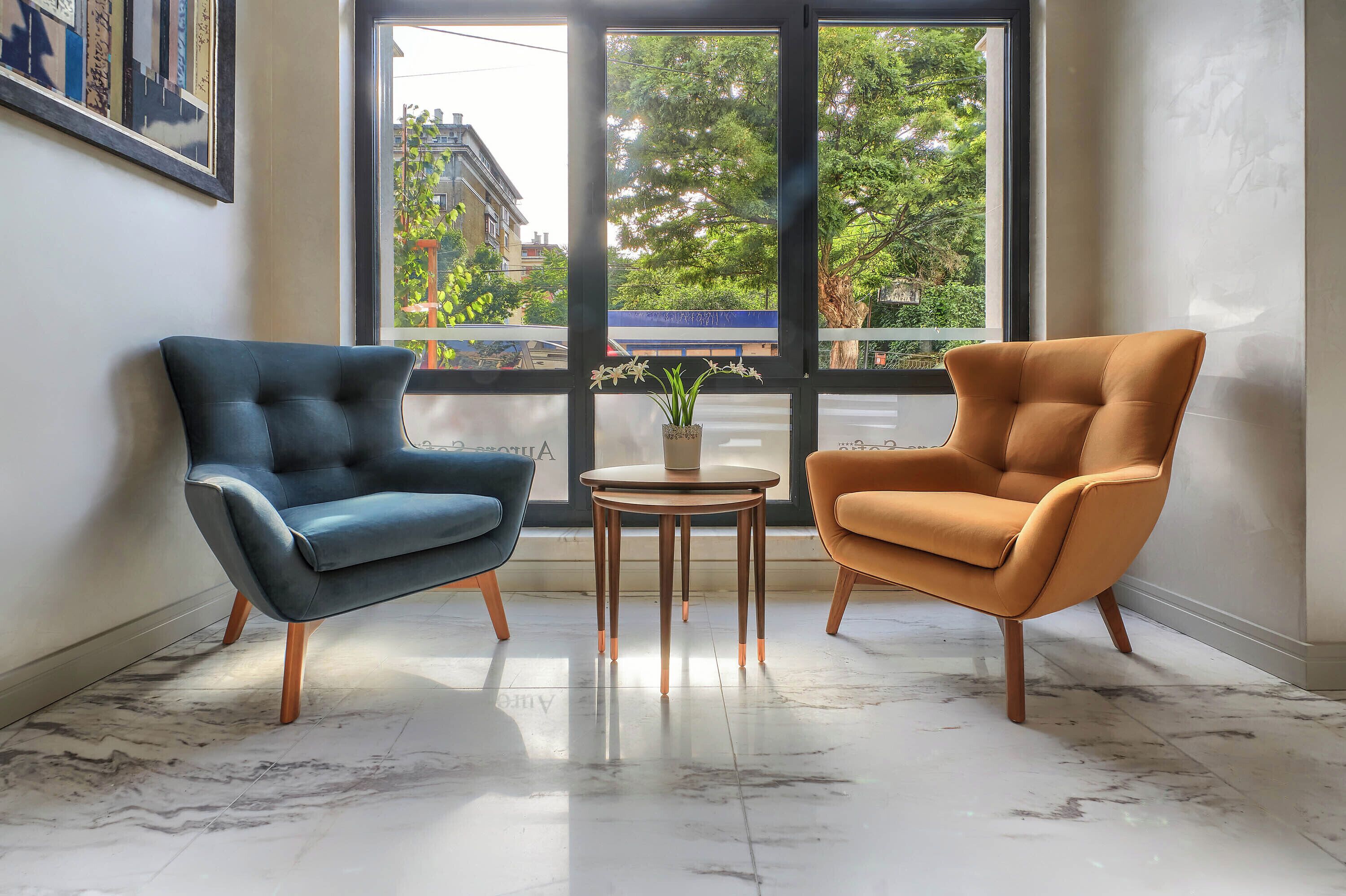 A room with two armchairs and a small table, a large window with a view of buildings and trees, and a marble floor.
