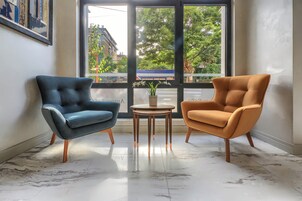 A room with two armchairs and a small table, a large window with a view of buildings and trees, and a marble floor.