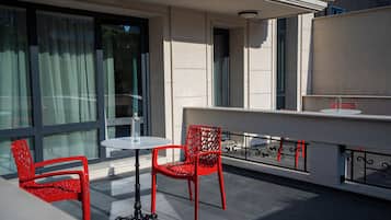 A terrace with two red chairs and a small round table, a glass balustrade, and a building with large windows and a flat roof.