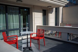 A terrace with two red chairs and a small round table, a glass balustrade, and a building with large windows and a flat roof.