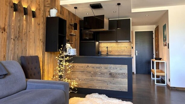 Un salon moderne avec un canapé gris, un mur d’accent en bois et un coin cuisine doté d’un pare-feu en bois et de meubles de rangement noirs.