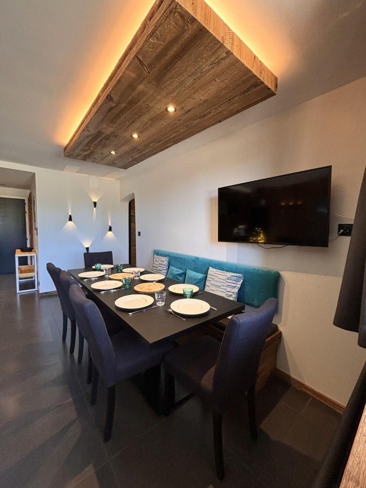 A modern dining area with a wooden ceiling, a flat-screen TV mounted on the wall, and a blue sofa.