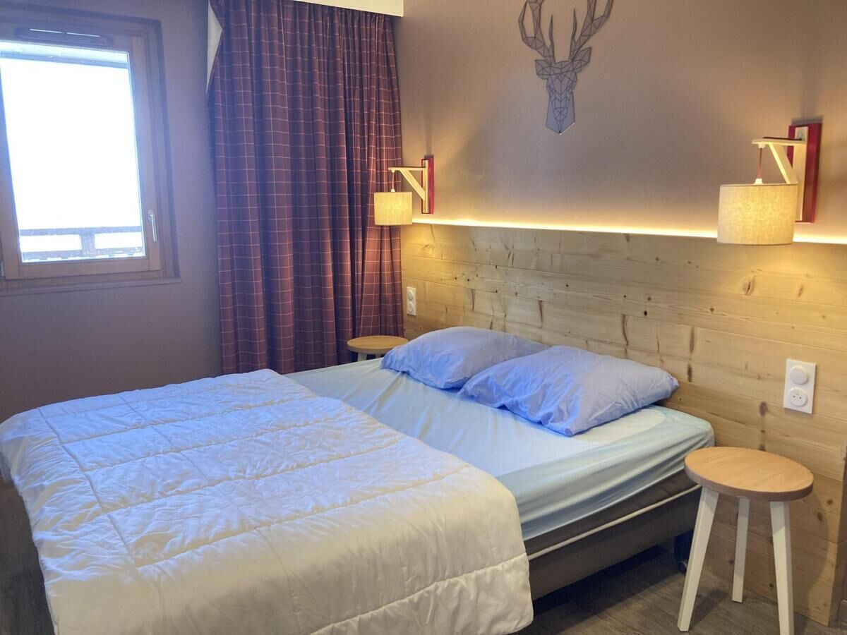 Een net opgemaakt bed met een witte deken en kussens, een houten hoofdbord, een wandlamp en een klein houten krukje.