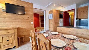 Comedor de madera con una mesa preparada para cuatro, un televisor montado en la pared y una cocina visible a través de una puerta abierta.