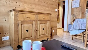 Un interior de madera con una mesa de comedor dispuesta con tazas blancas sobre un mantel rojo con lunares, un televisor montado en la pared y un mueble de madera con recortes en forma de corazón.