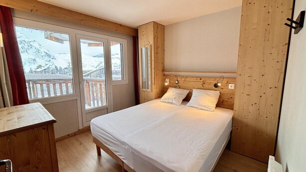 Un dormitorio con cama, cabecero de madera y vistas a montañas nevadas a través de amplios ventanales.