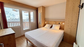 Quarto com cama, cabeceira de madeira e vista para montanhas nevadas através de grandes janelas.