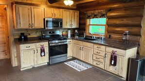 A log cabin kitchen with wooden cabinets, stainless steel appliances, and a window with a view of greenery. - Up North Retreat near Lake Superior (Port Wing)