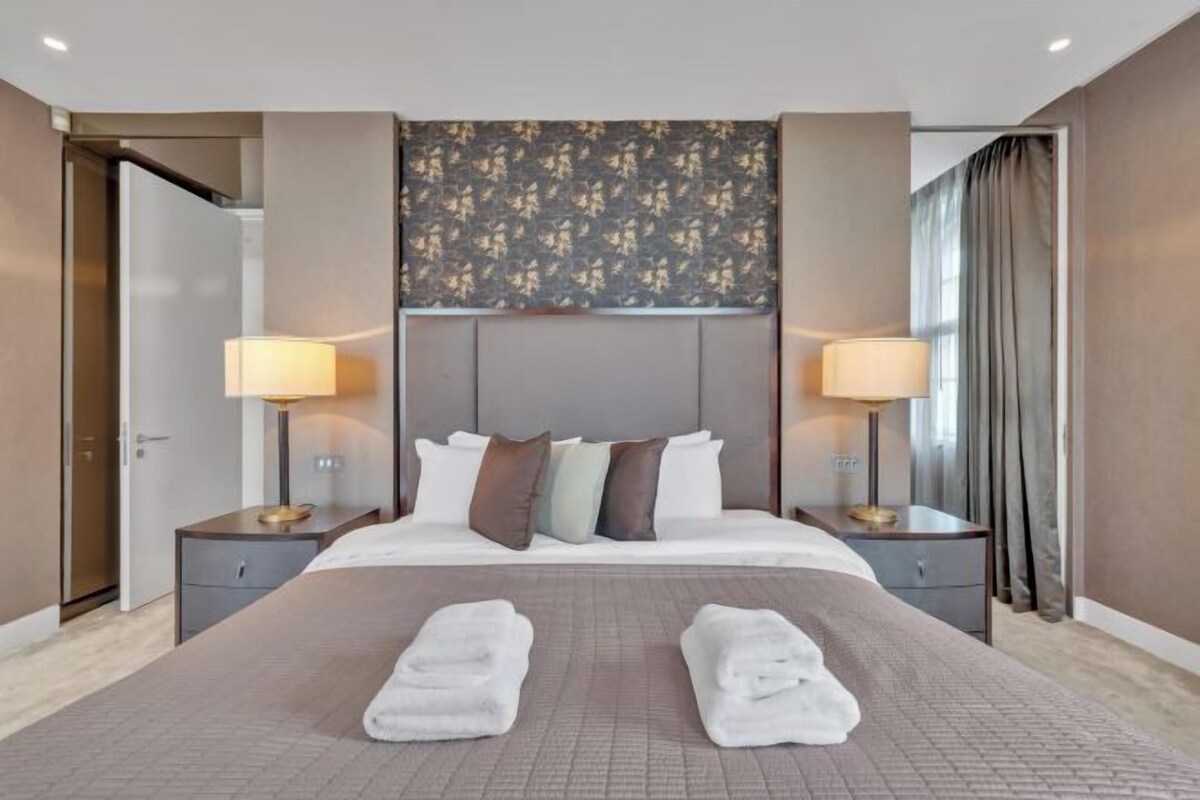 A hotel room with a large bed, two bedside tables, a headboard with a floral pattern, and a view of the outside through a window.