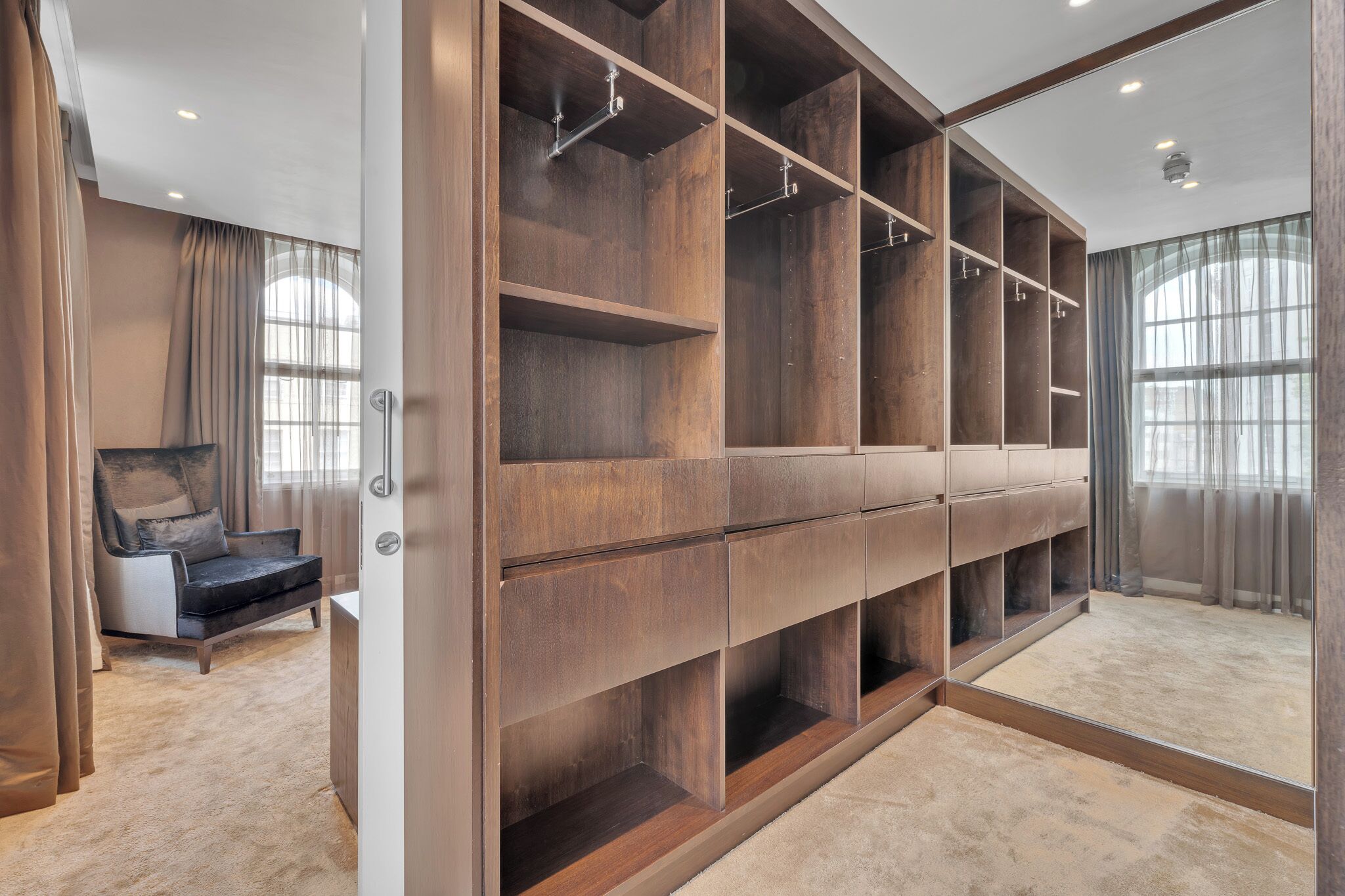 Ein begehbarer Kleiderschrank mit zahlreichen Regalen und Ablageflächen, ein Sessel sowie große Fenster mit Vorhängen.