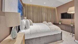A modern bedroom with a large bed, a bedside table with a lamp, a wall-mounted TV, and a window with curtains.