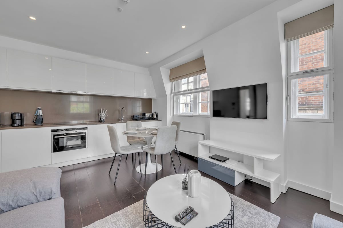 A modern kitchen with white cabinets, a dining area with a round table, and a TV mounted on the wall.