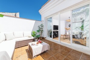 Un balcón moderno con un sofá blanco, una mesita con una planta y una puerta corrediza de vidrio que da a un espacio interior.