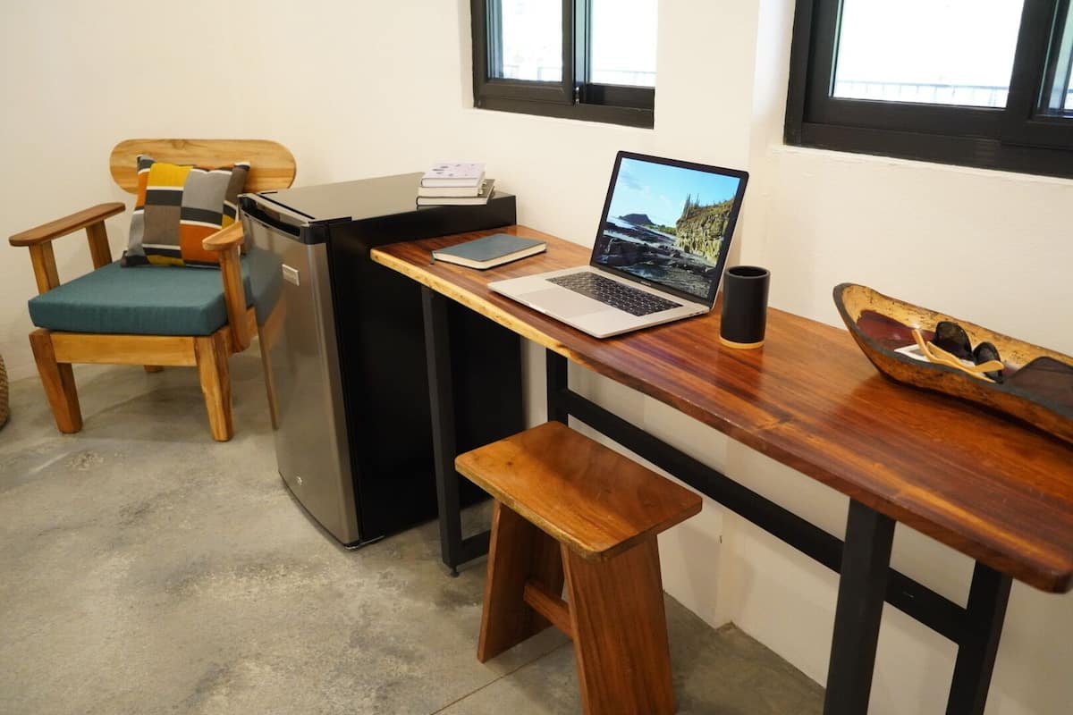 A compact kitchenette with a wooden desk, a laptop, a refrigerator, a bench, and a small table with a decorative bowl.