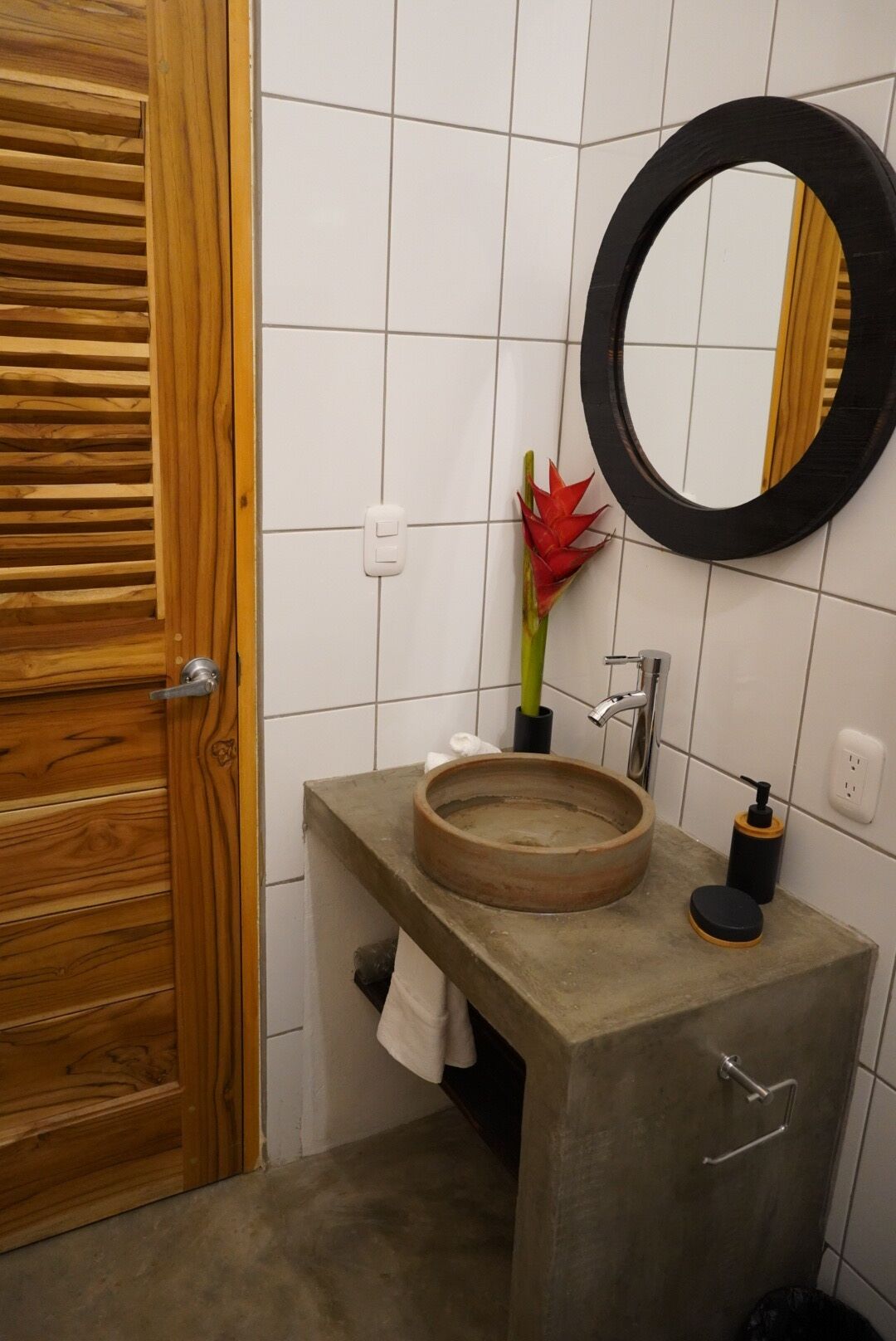 Un baño con puerta de madera, un lavabo redondo con cuenca de madera, un espejo negro y una flor roja en un florero.