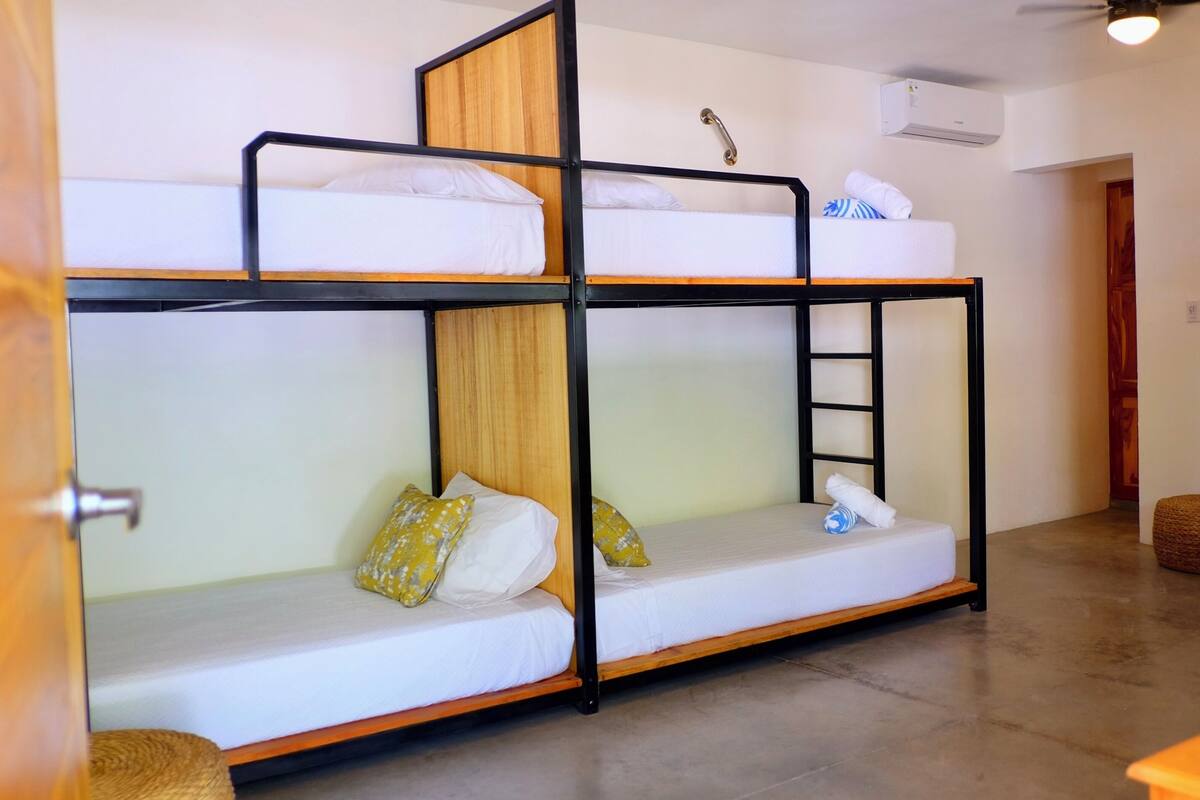 A bunk bed room with a wooden ladder, white bedding, and a woven ottoman.