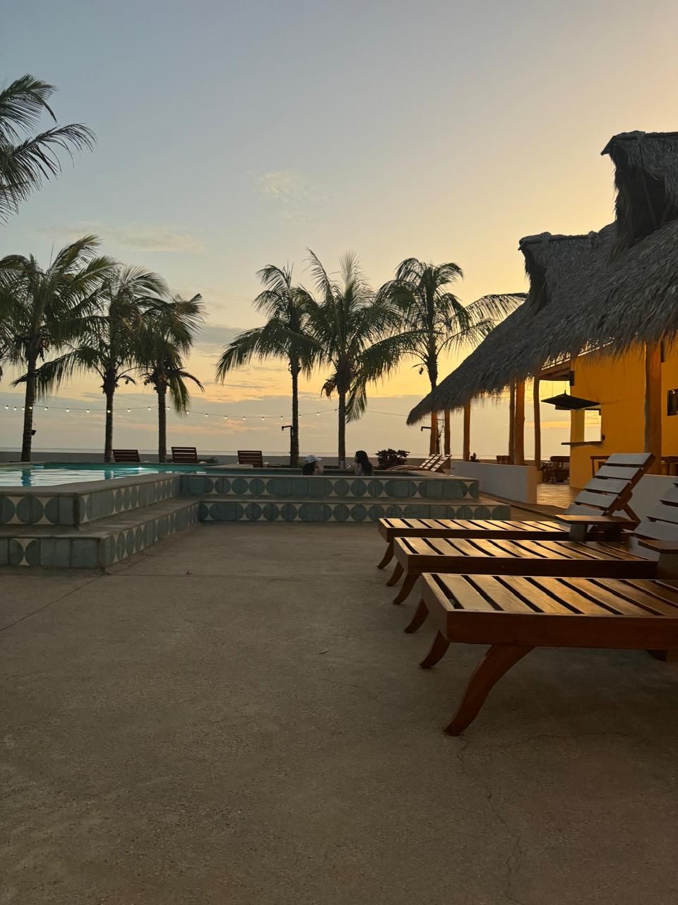 Ein Poolbereich mit Liegestühlen und einer strohgedeckten Struktur, mit Blick auf einen Strand.