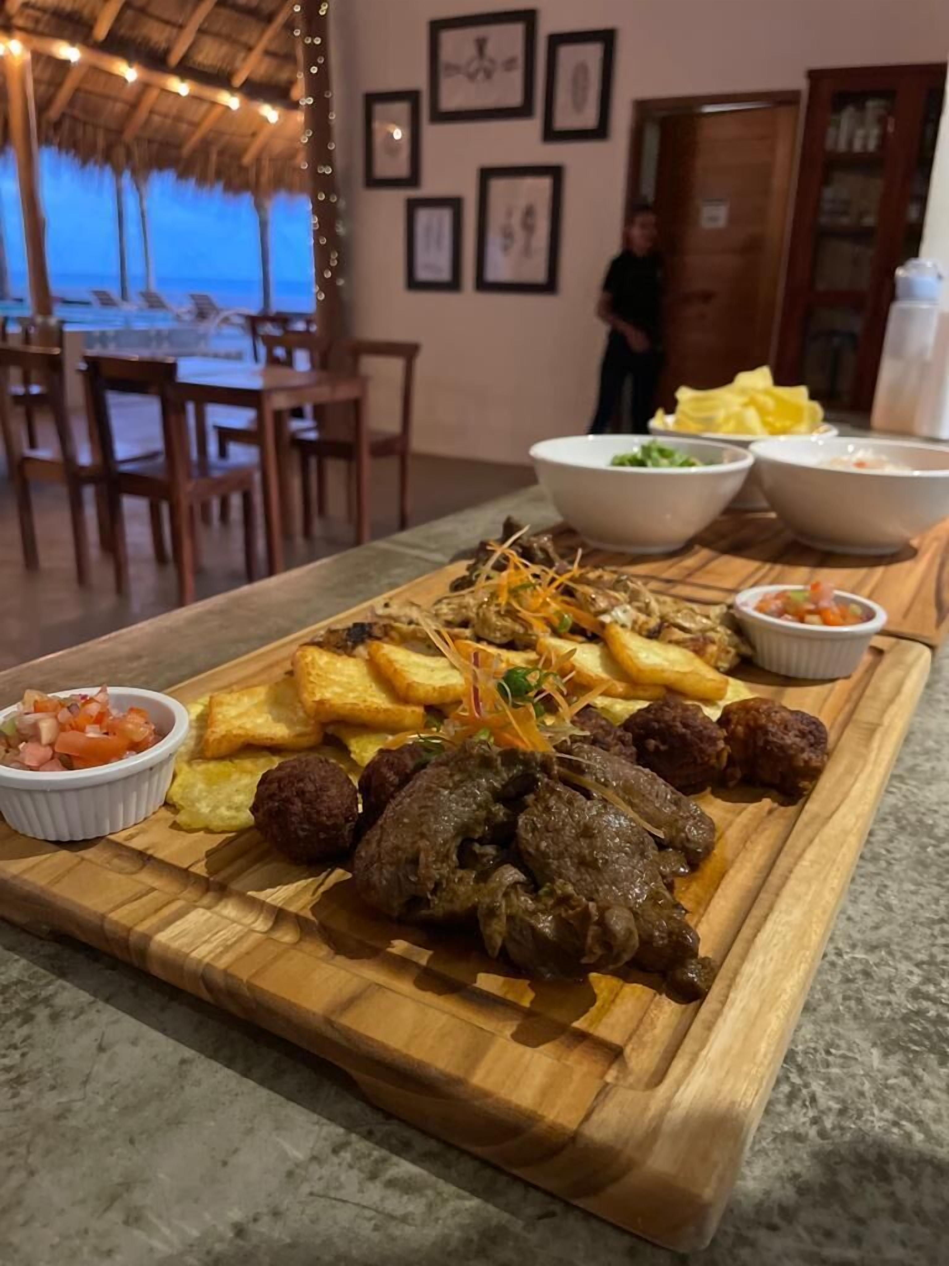 Una tabla de madera con diversos platos, incluyendo carne, papas fritas y salsas, colocada en una habitación con techo de paja y cuadros en la pared.