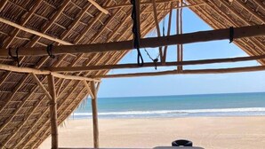 A room with a wooden floor and a bed with a white sheet. The room has a thatched roof and a view of the beach and ocean. - Hotel Kraken (Tola)