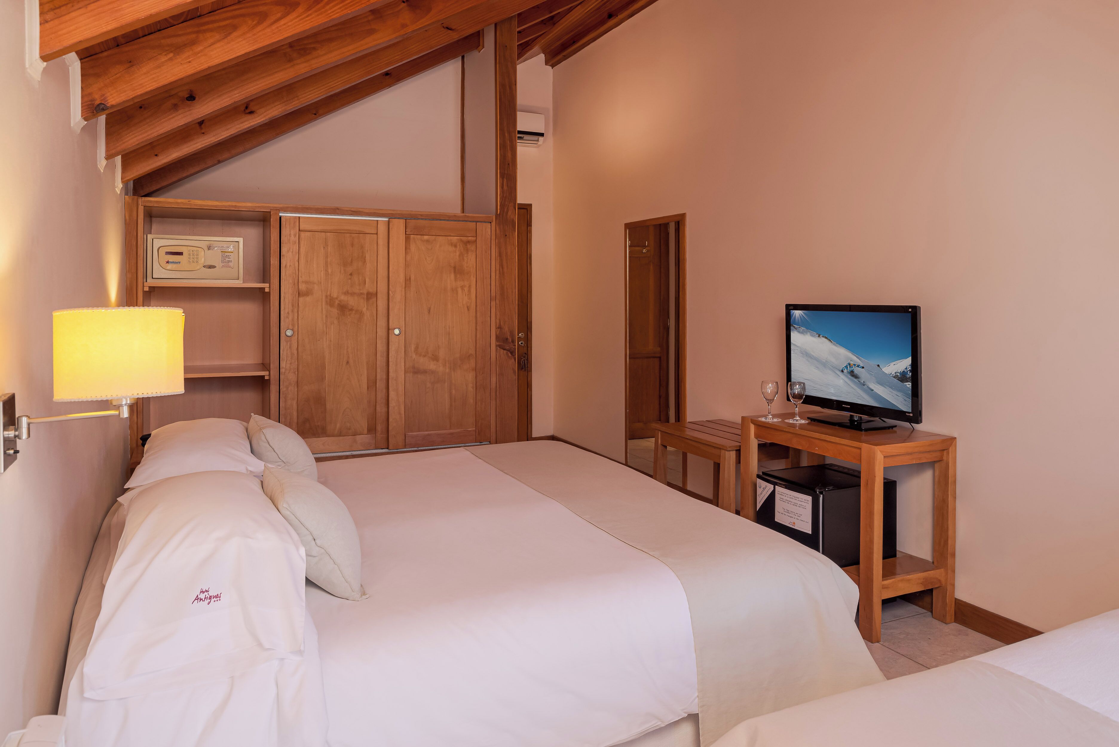 Quarto de hotel com uma cama, televisão sobre um suporte de madeira, escrivaninha com abajur e guarda-roupa.