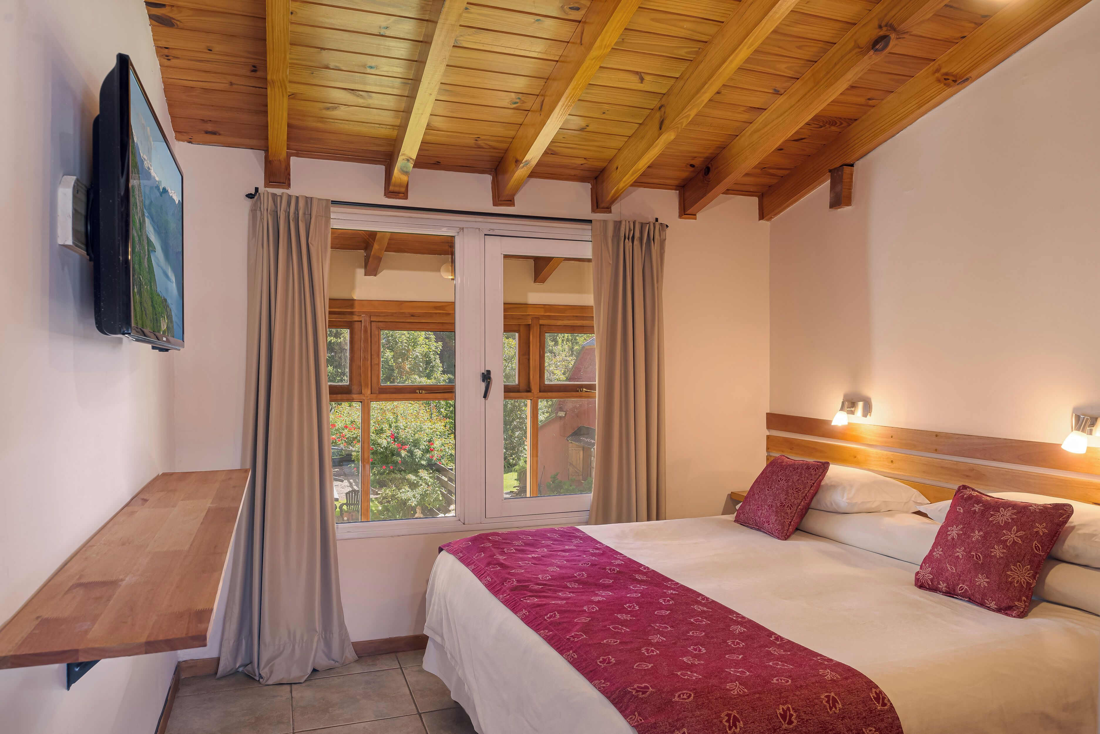 A bedroom with a wooden ceiling, a bed with a red patterned bedspread, a wooden nightstand, a TV mounted on the wall, and a window with curtains.
